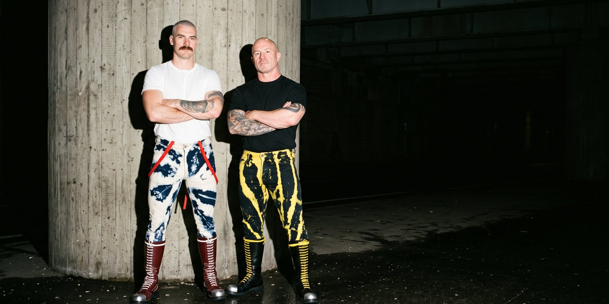 Two men under a flyover at night in hand-bleached jeans, one pair blue and white with red braces, one black and yellow, with tall lace-up boots.